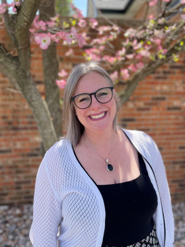 Chelsea France, Director of Client Relations at Inline Data Systems, wears a white cardgan and black undershirt in front of a brick wall and tree.